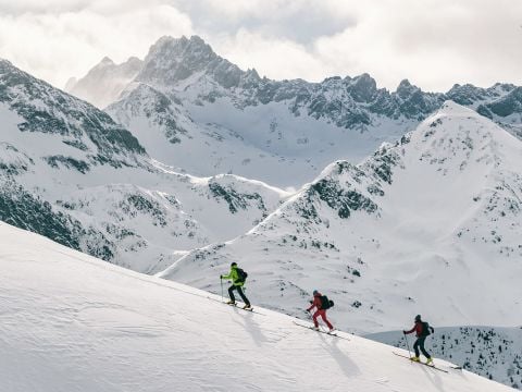 Drei Skifahrer in farbenfrohen Outfits fahren einen verschneiten Berghang hinauf, vor der Kulisse schroffer, schneebedeckter Gipfel. Der Himmel ist bedeckt, ein Hauch von Sonnenlicht bricht durch und wirft ein sanftes Licht auf die Szene.