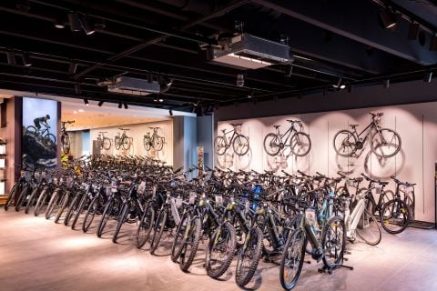 Ein modernes Interieur eines Fahrradladens mit Reihen von Fahrrädern auf dem Boden und mehreren an den Wänden. Der Raum ist gut beleuchtet und verfügt über glatte, dunkle Decken und stimmungsvolles Licht. Bilder von Radfahrern schmücken die Wände und verstärken die sportliche Atmosphäre.